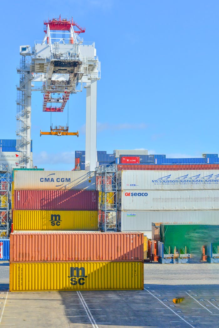 our-journey-01 Stacked cargo containers in a sunny port industrial setting with crane.