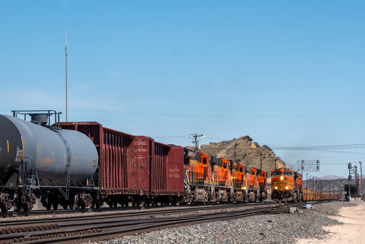 services-02 Orange and red freight train traveling on desert railway tracks under a clear blue sky.