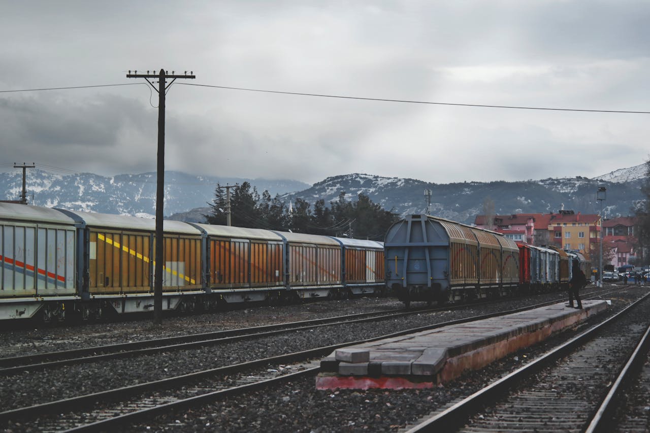 who-we-are-img A freight train with colorful cargo wagons on a mountain railway track under a gray sky.